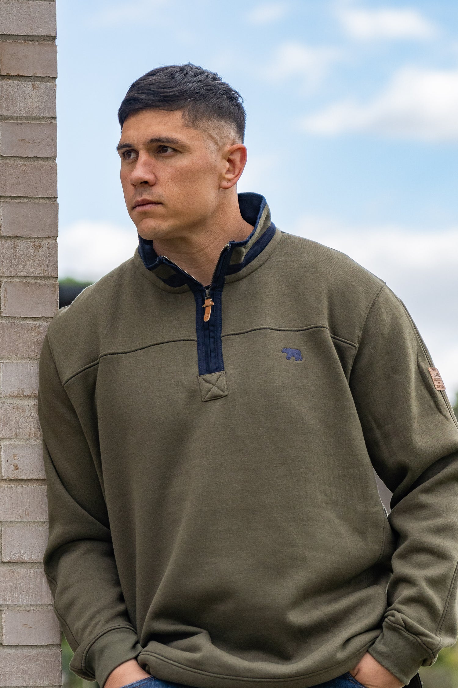 Man wearing a green pullover with a logo, standing against a brick wall with a blue sky.