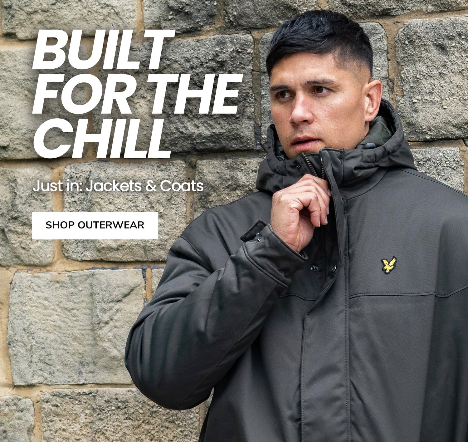 Man wearing a green jacket with a lyle and scott logo against a stone wall, promoting jackets and coats