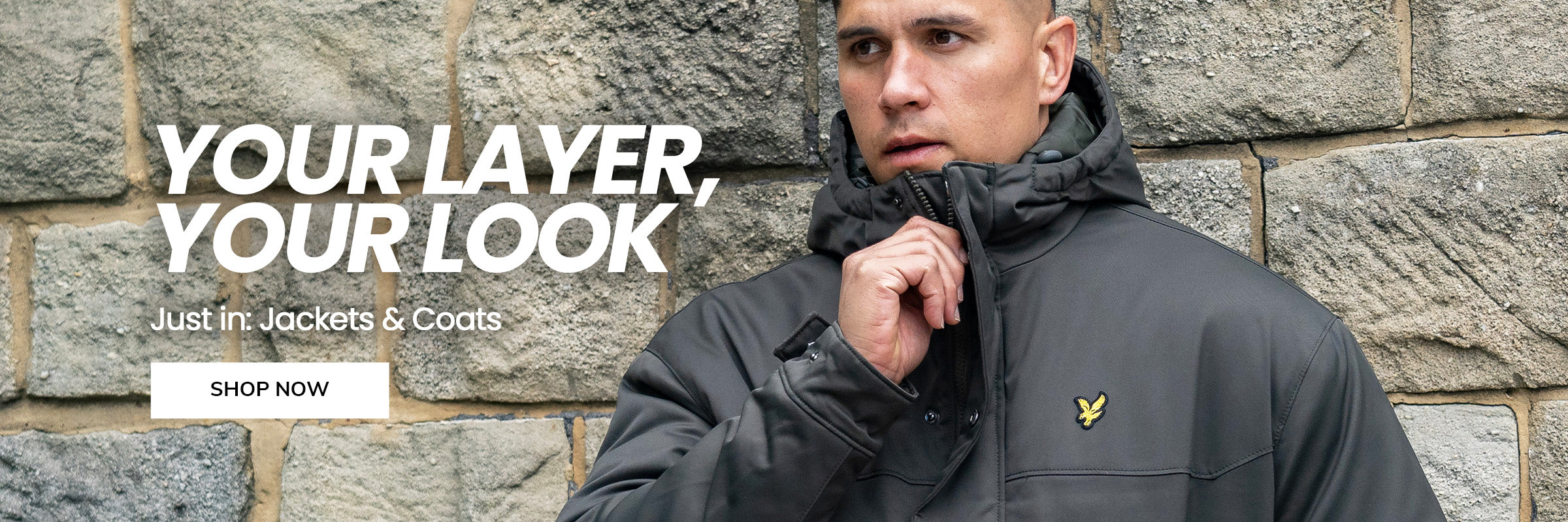 Man wearing a green jacket with a lyle and scott logo against a stone wall, promoting jackets and coats
